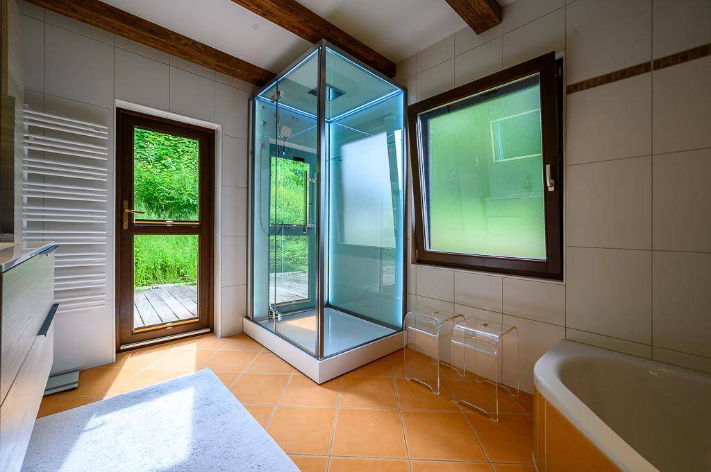 Helles Badezimmer mit Duschkabine, Badewanne und Fenster mit Blick ins Grüne.