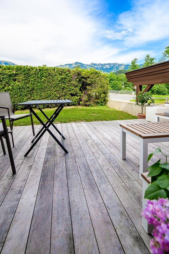Holzterrasse mit Gartenmöbeln, Hecke und Bergblick unter blauem Himmel.