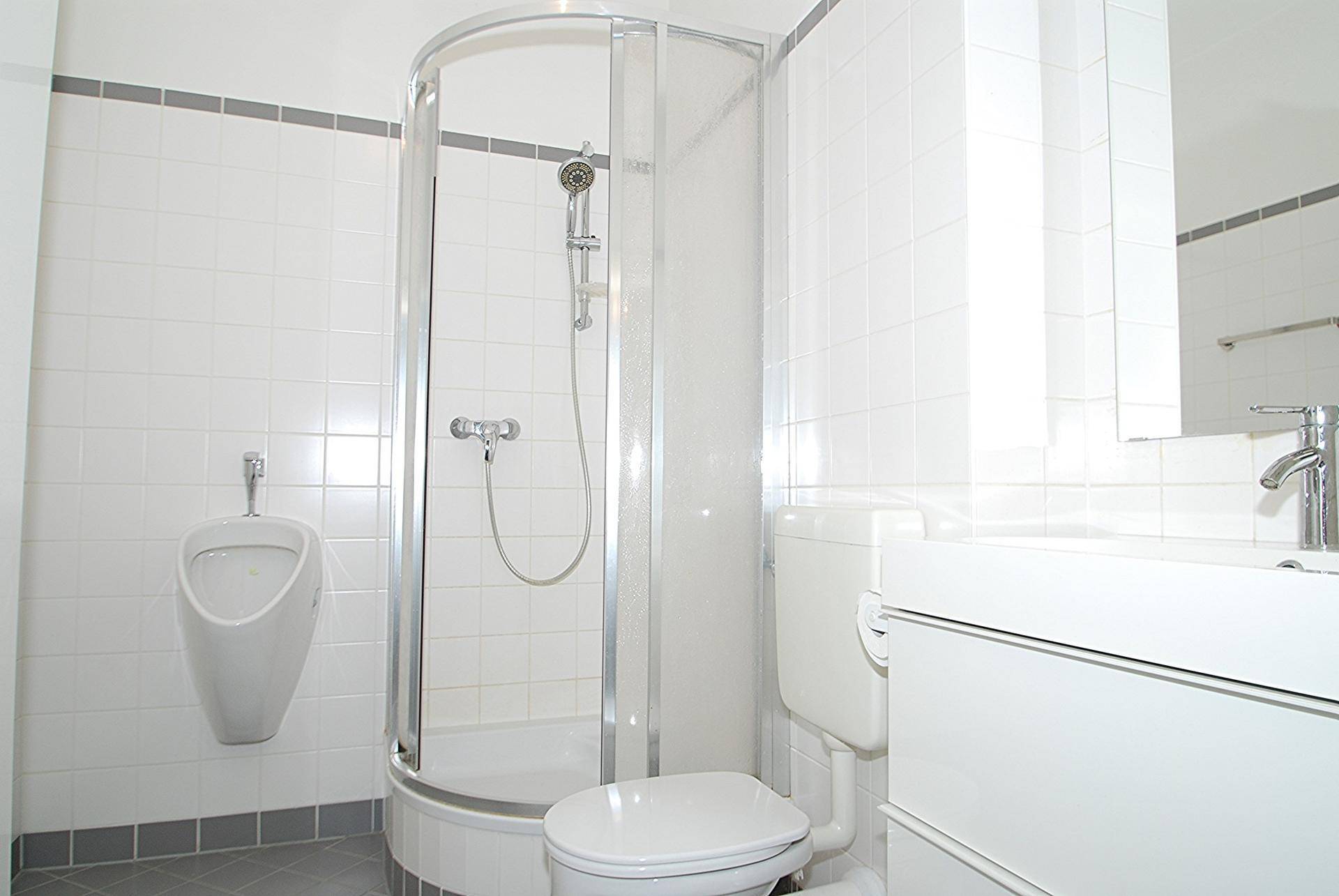 Helles Badezimmer mit Duschkabine, WC und Urinal, weiß gefliest.