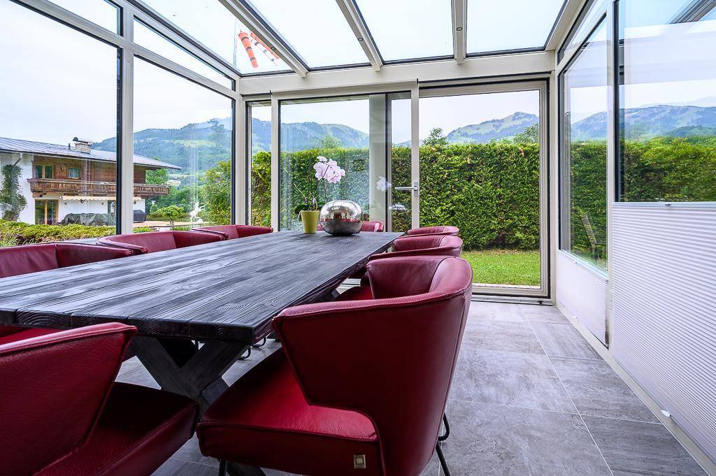 Heller Wintergarten mit großem Esstisch, roten Stühlen und weitem Blick auf die Berge.