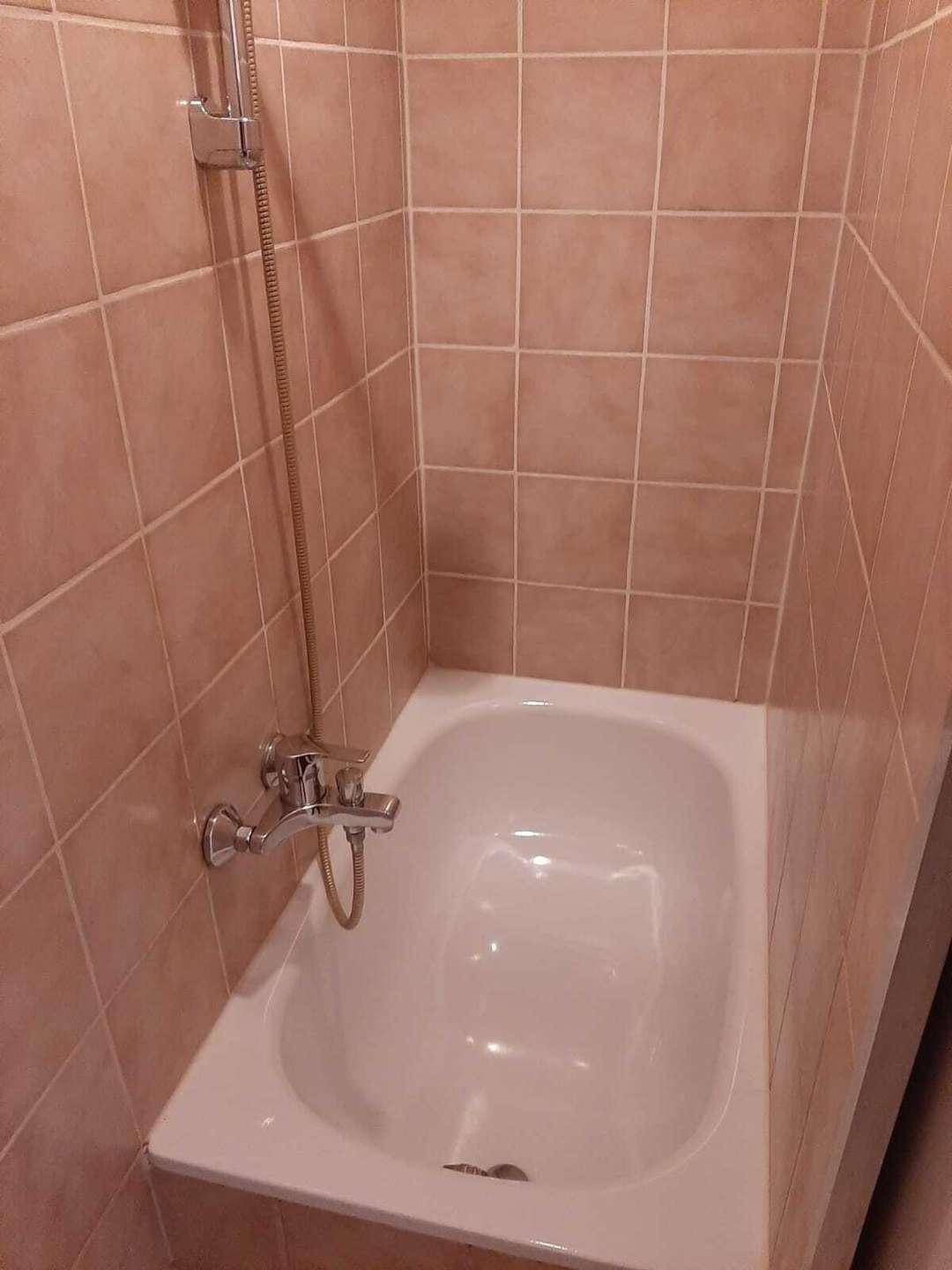 Kleines Badezimmer mit Badewanne und Duscharmatur, vollständig gefliest in hellem Braunton.