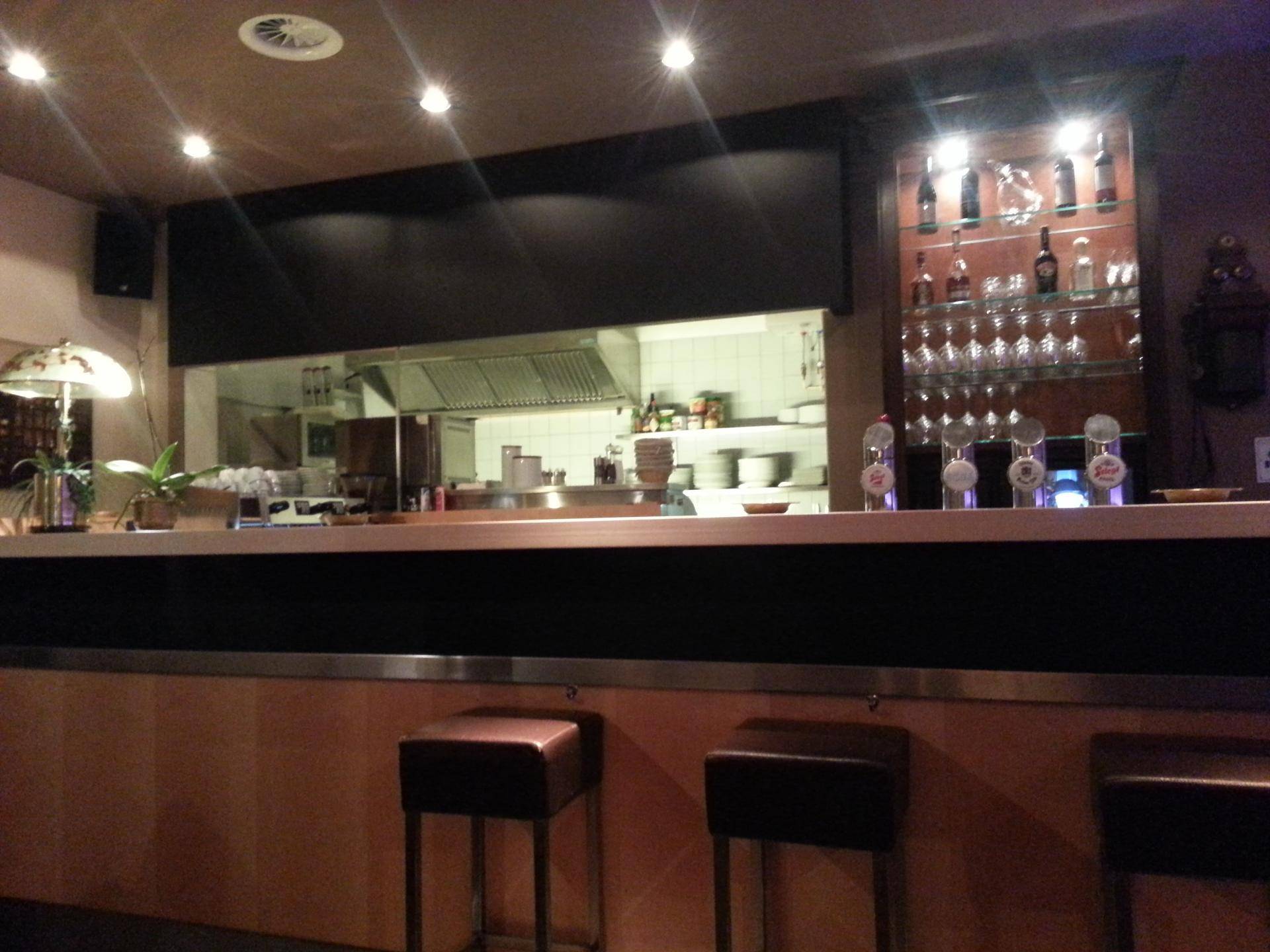 Blick auf die Bar und die offene Küche bei Nacht, mit beleuchteter Arbeitsfläche und Flaschenregal.