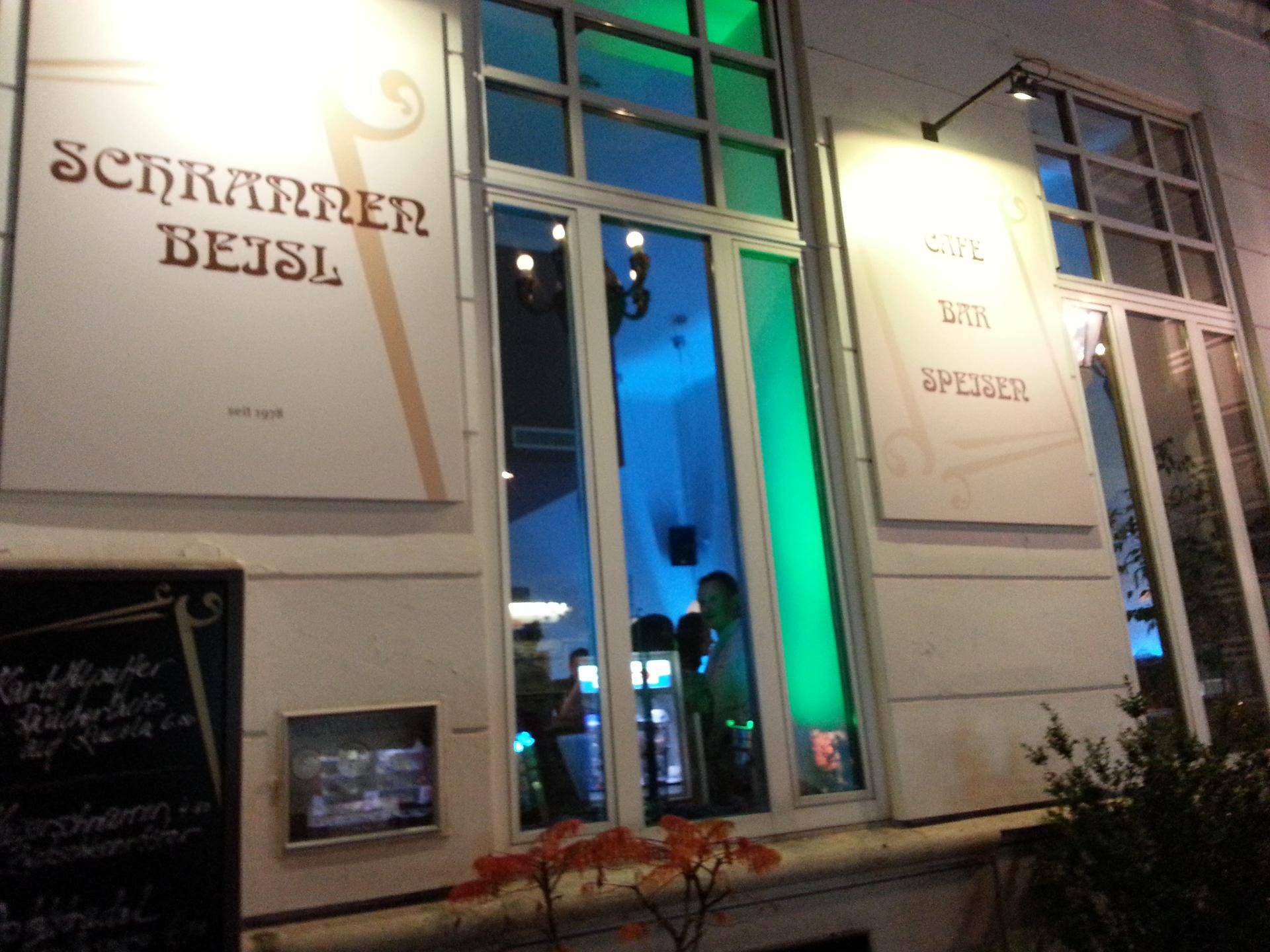 Fassade des Cafés bei Nacht, mit farbiger Innenbeleuchtung, die durch die Fenster scheint.