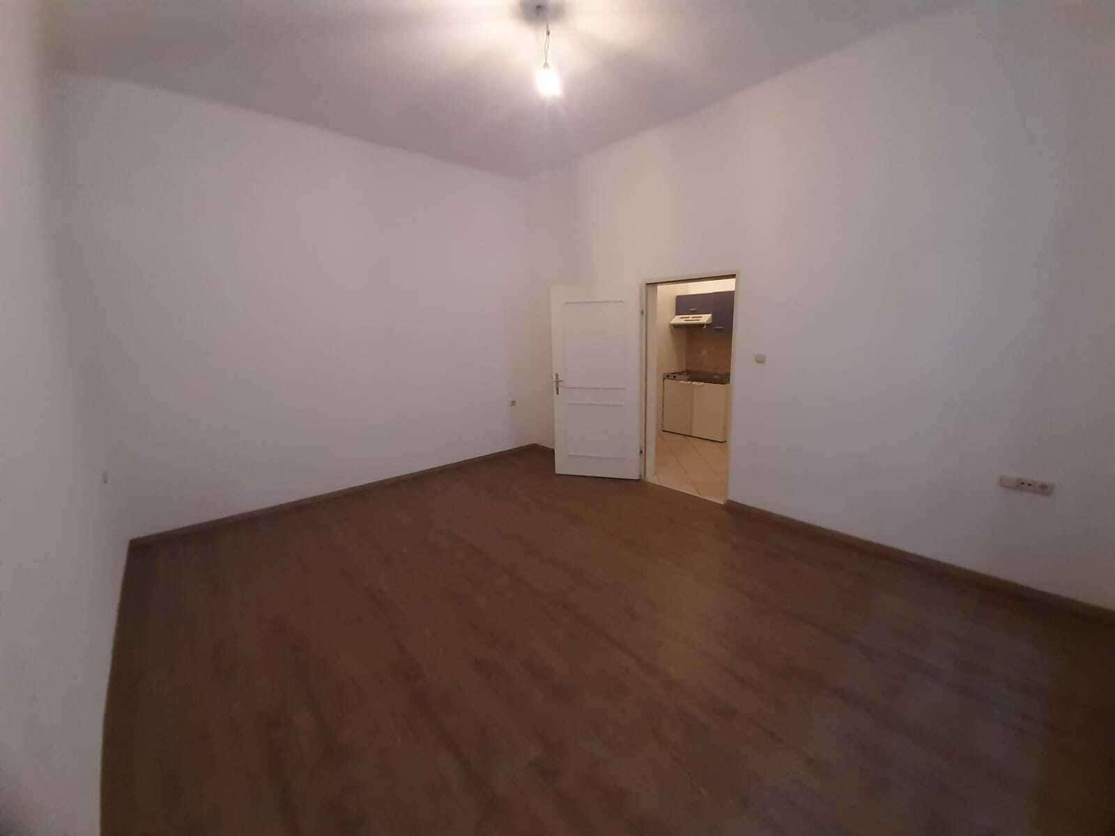 Leeres Zimmer mit Holzboden und Zugang zu einer kleinen Küche, ideal für Singles oder Paare.