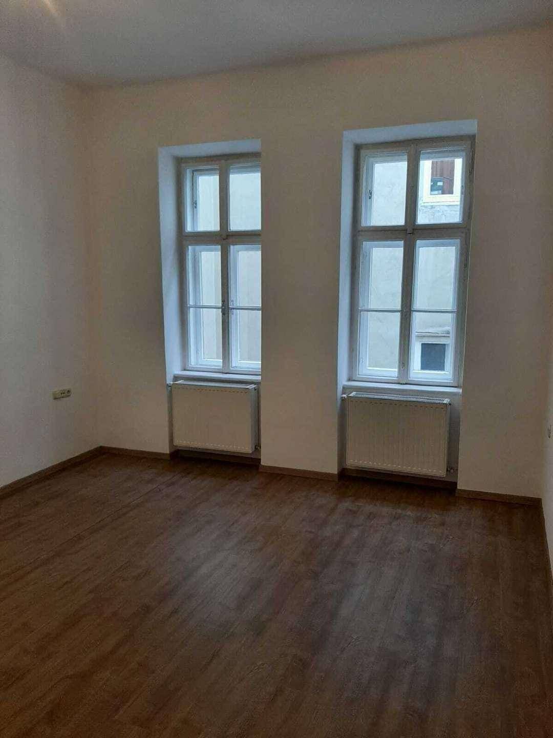 Unmöbliertes Zimmer mit zwei hohen Fenstern und Heizkörpern, bietet viel Tageslicht.