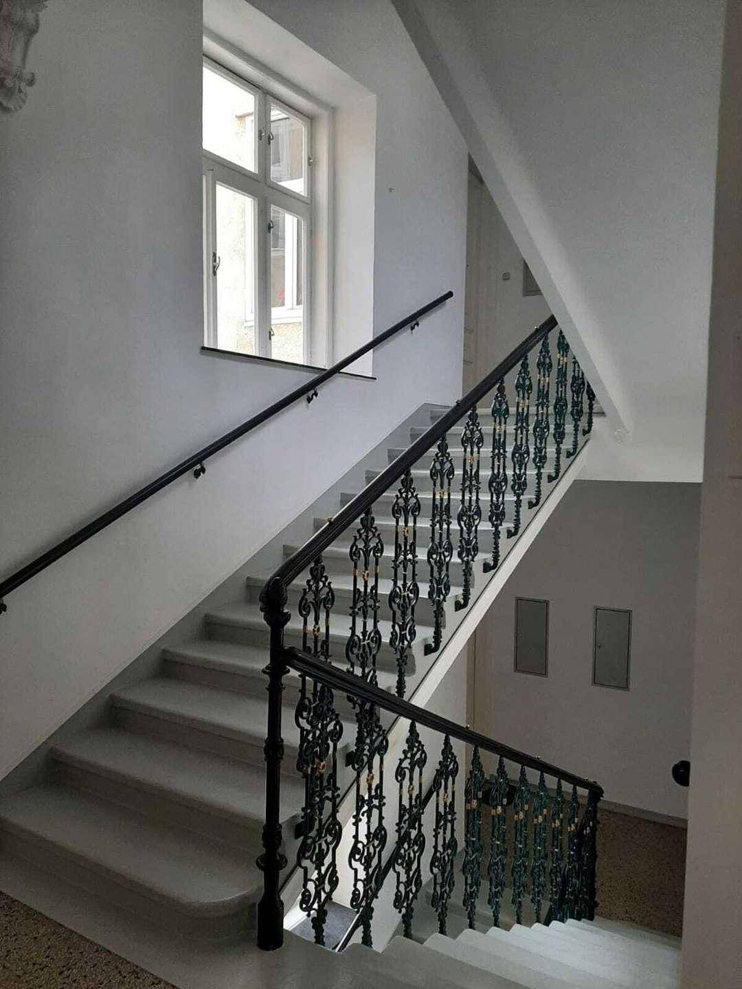 Historisches Treppenhaus mit kunstvollem schmiedeeisernem Geländer und hohen Fenstern.