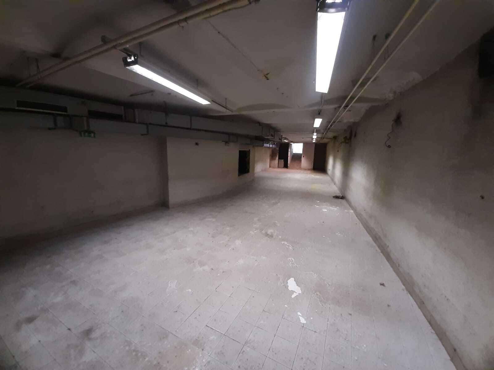 Großer, leerer Gewerberaum im Kellergeschoss mit Fliesenboden und sichtbaren Lüftungsrohren.