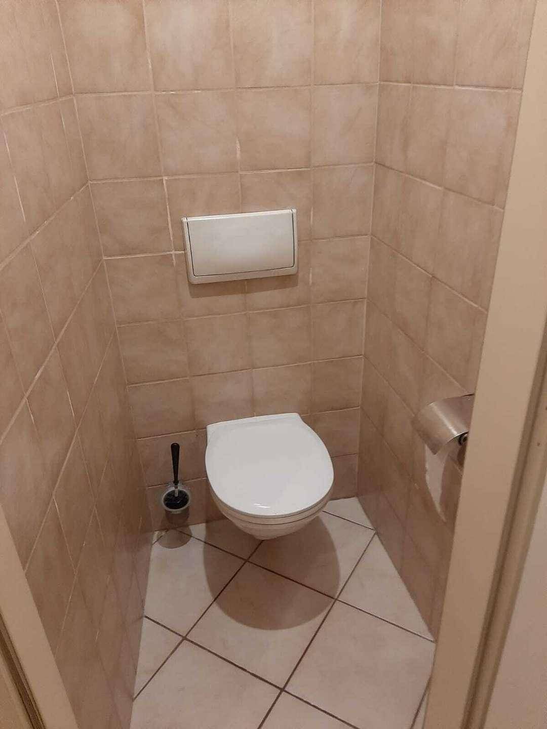 Separates WC mit wandmontierter Toilette und hellbraunen Fliesen an Wänden und Boden.