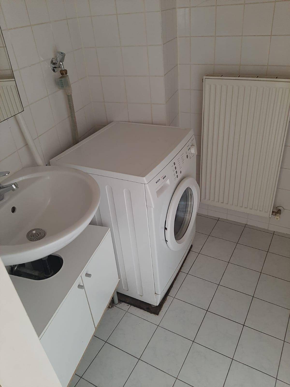 Badezimmer mit Waschbecken, Waschmaschine und weißen Wand- und Bodenfliesen, inklusive Heizkörper.