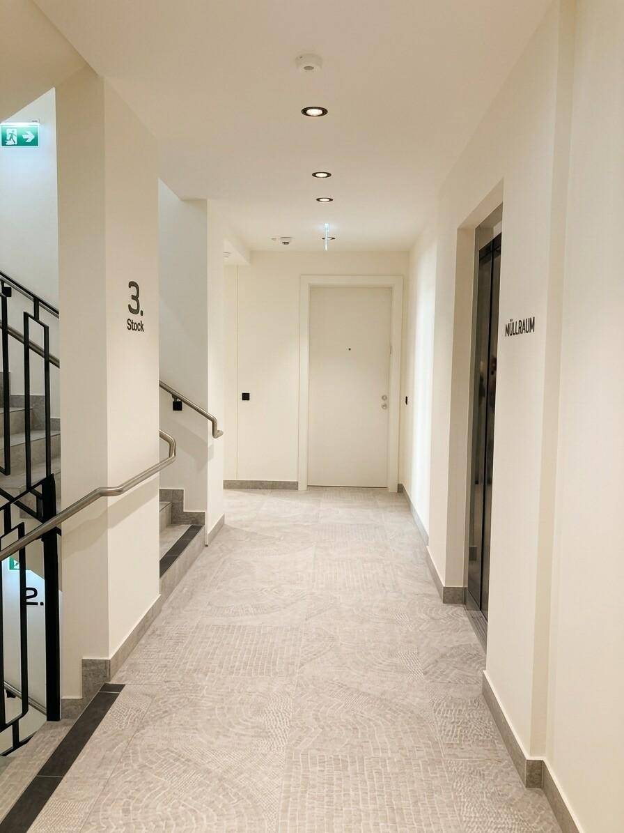 Heller Flur im Gebäude mit Treppenhaus, Aufzug und Zugang zu den Wohnungen.