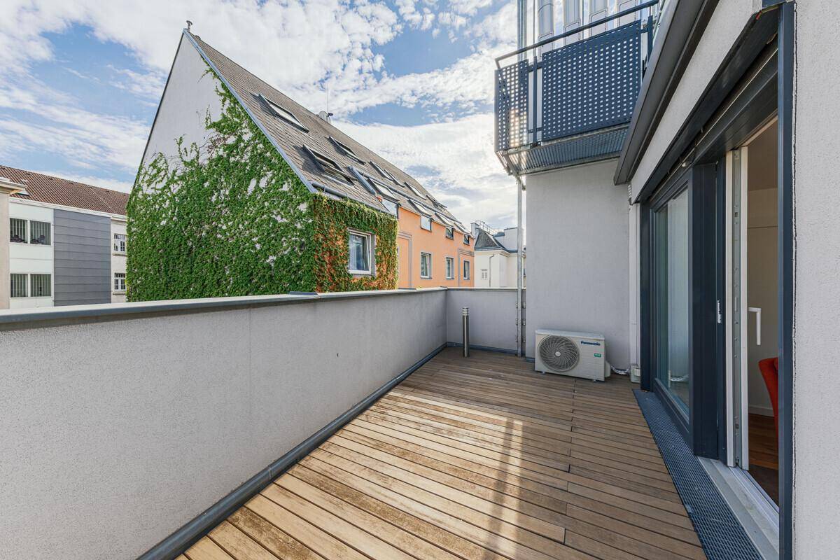 Balkon mit Holzbelag, Klimaanlage und Blick auf ein begrüntes Gebäude in der Nachbarschaft.