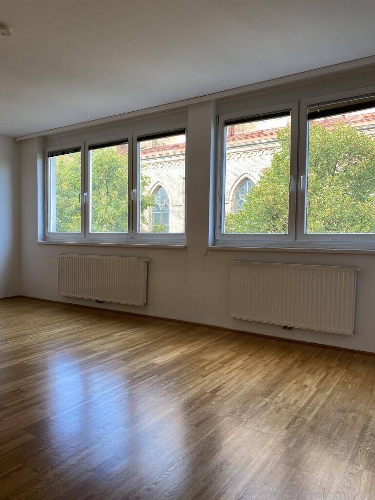 Heller Raum mit Parkettboden und großen Fenstern, die einen Blick auf ein historisches Gebäude bieten.