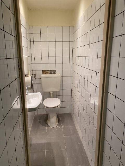 Kleine Toilettenkabine mit weißen Wandfliesen, WC und Waschbecken, einfach ausgestattet und renovierungsbedürftig.
