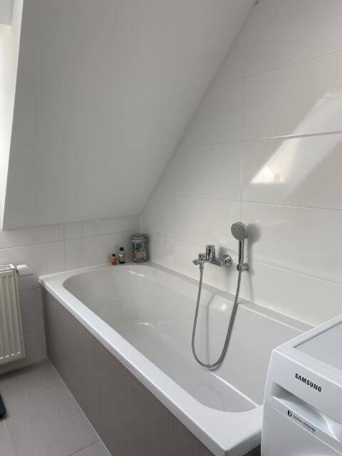 Helle Badewanne in einem gefliesten Badezimmer mit Dachschräge und Waschmaschine.