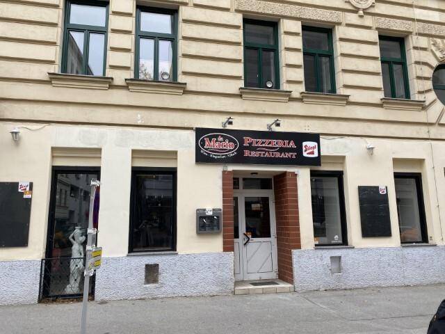 Fassade eines Gebäudes mit dem Eingang zu einem Restaurant, gekennzeichnet durch grüne Fensterrahmen und eine helle Außenwand.
