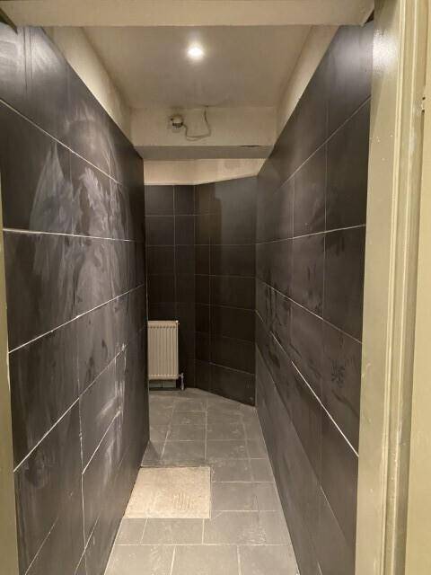 Langer Flur oder Umkleidebereich mit dunklen Fliesen an Wänden und Boden, Heizkörper sichtbar, renovierungsbedürftig.