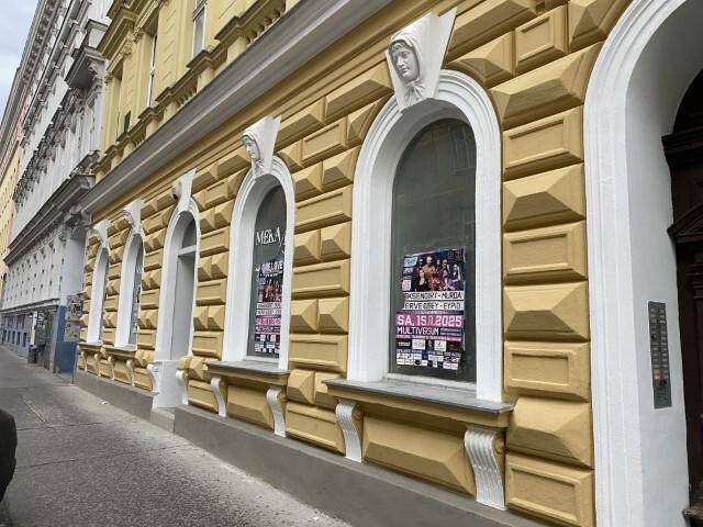 Historische Gebäudefassade in Gelb mit dekorativen Bögen und Werbeplakaten an den Fenstern im Erdgeschoss.