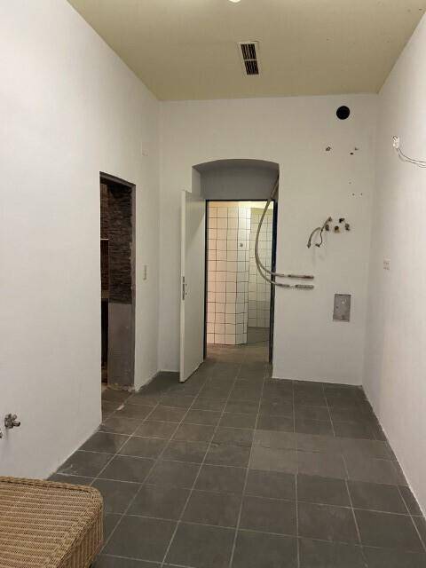 Leerer Raum mit grauen Fliesen und sichtbaren Wasseranschlüssen, der zu einem Duschbereich führt, renovierungsbedürftig.