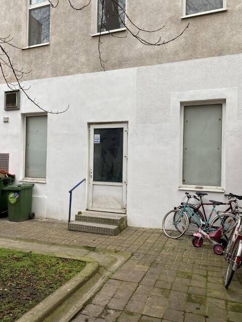 Weiße Gebäudefassade mit Eingangstür und Fenstern, Fahrräder stehen vor dem Gebäude auf dem Gehweg.