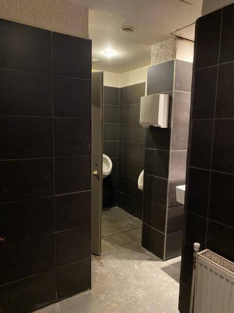 Herren-Waschraum mit Urinalen, Waschbecken und dunklen Fliesen, funktional eingerichtet und renovierungsbedürftig.