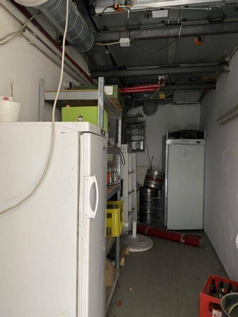 Lagerraum im Keller mit Regalen, Kühlschränken und sichtbaren Leitungen, genutzt für Getränke und Vorräte.