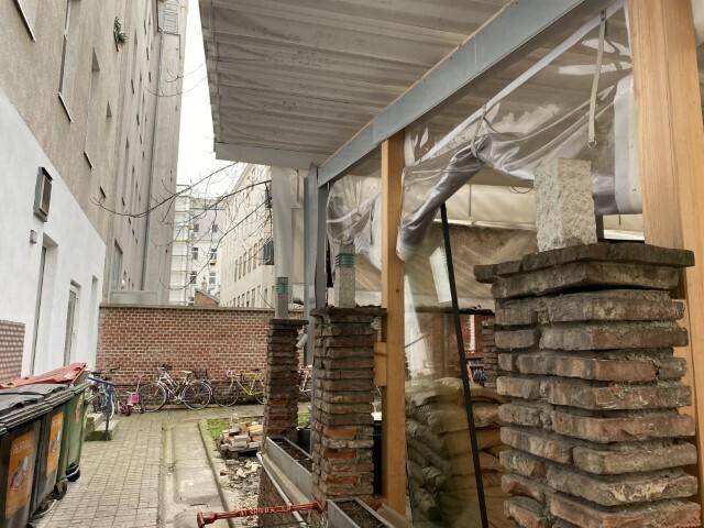 Baustelle mit freiliegenden Ziegelpfeilern und provisorischer Überdachung, umgeben von Gebäuden.