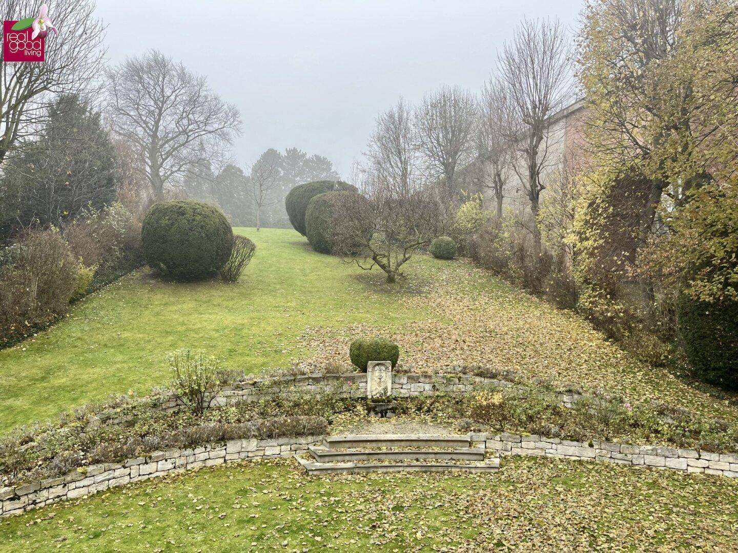 Herbstliche Gartenlandschaft mit terrassierten Beeten, alten Bäumen und einer Steintreppe in nebliger Atmosphäre.
