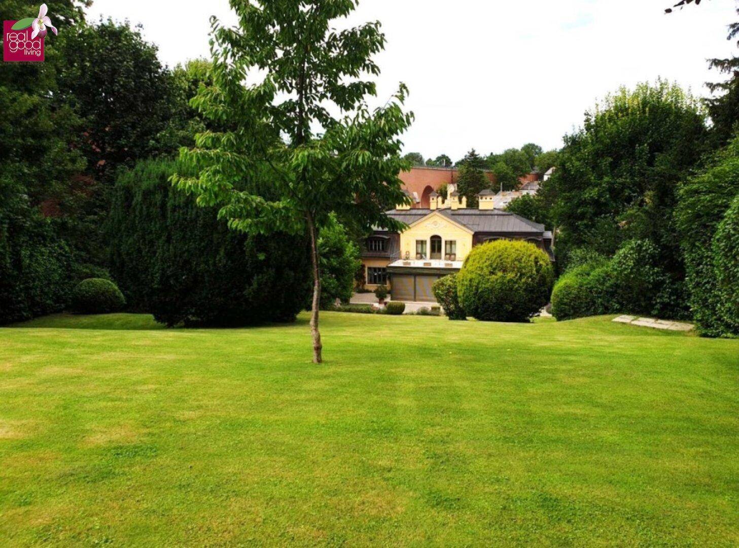 Große Rasenfläche im weitläufigen Garten mit Blick auf die Villa und umgebende Bäume.