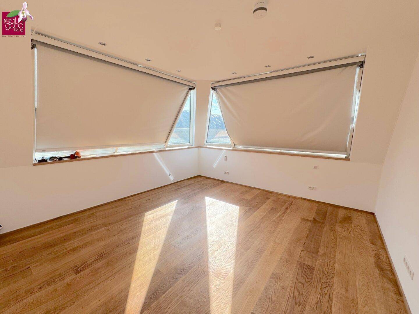 Heller Raum mit Holzboden, schrägen Wänden und zwei Fenstern mit Rollos, ideal als Schlafzimmer.