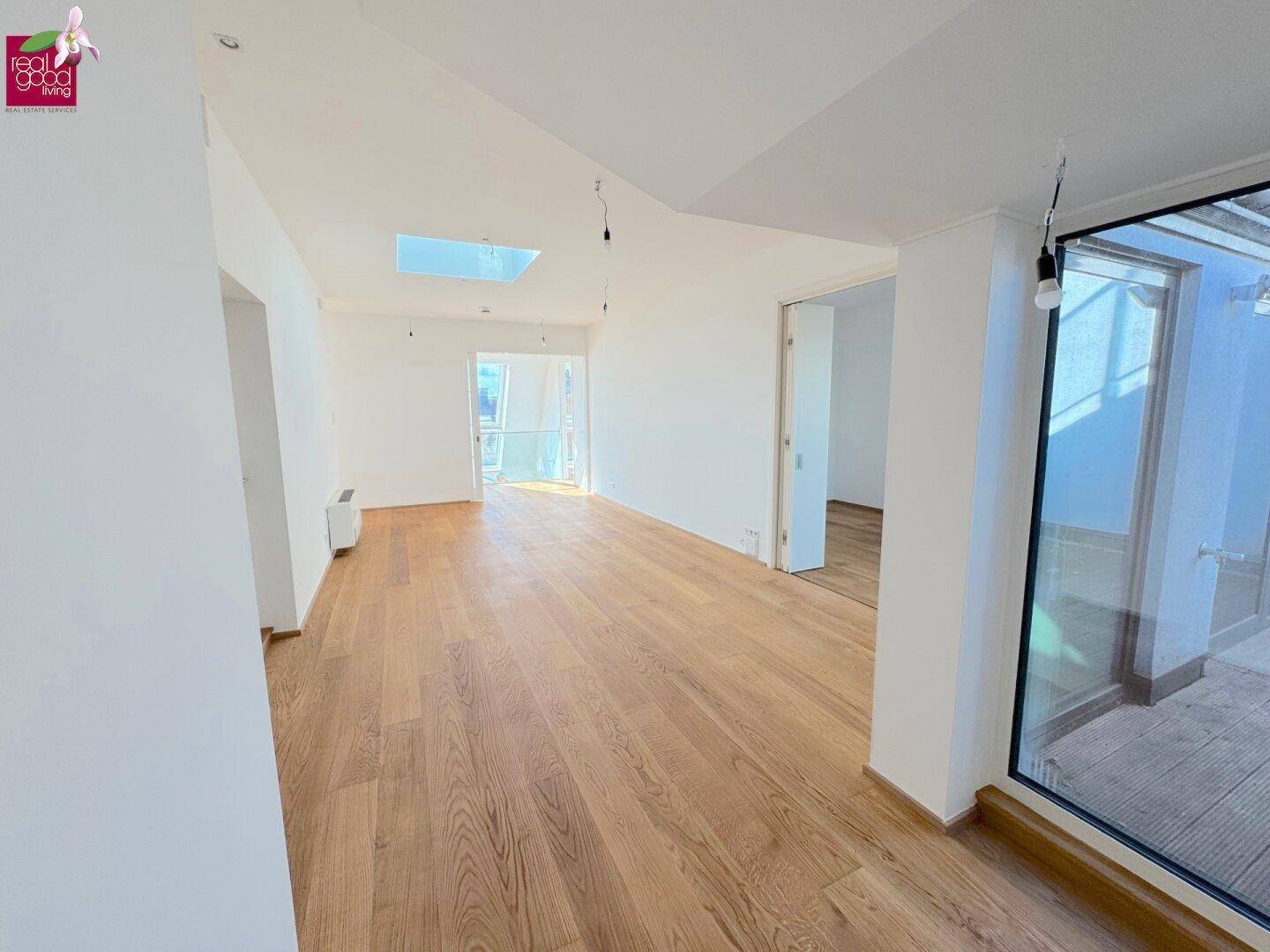 Heller Wohnbereich mit Holzboden, Oberlicht und Zugang zu einem Außenbereich durch große Fenster.