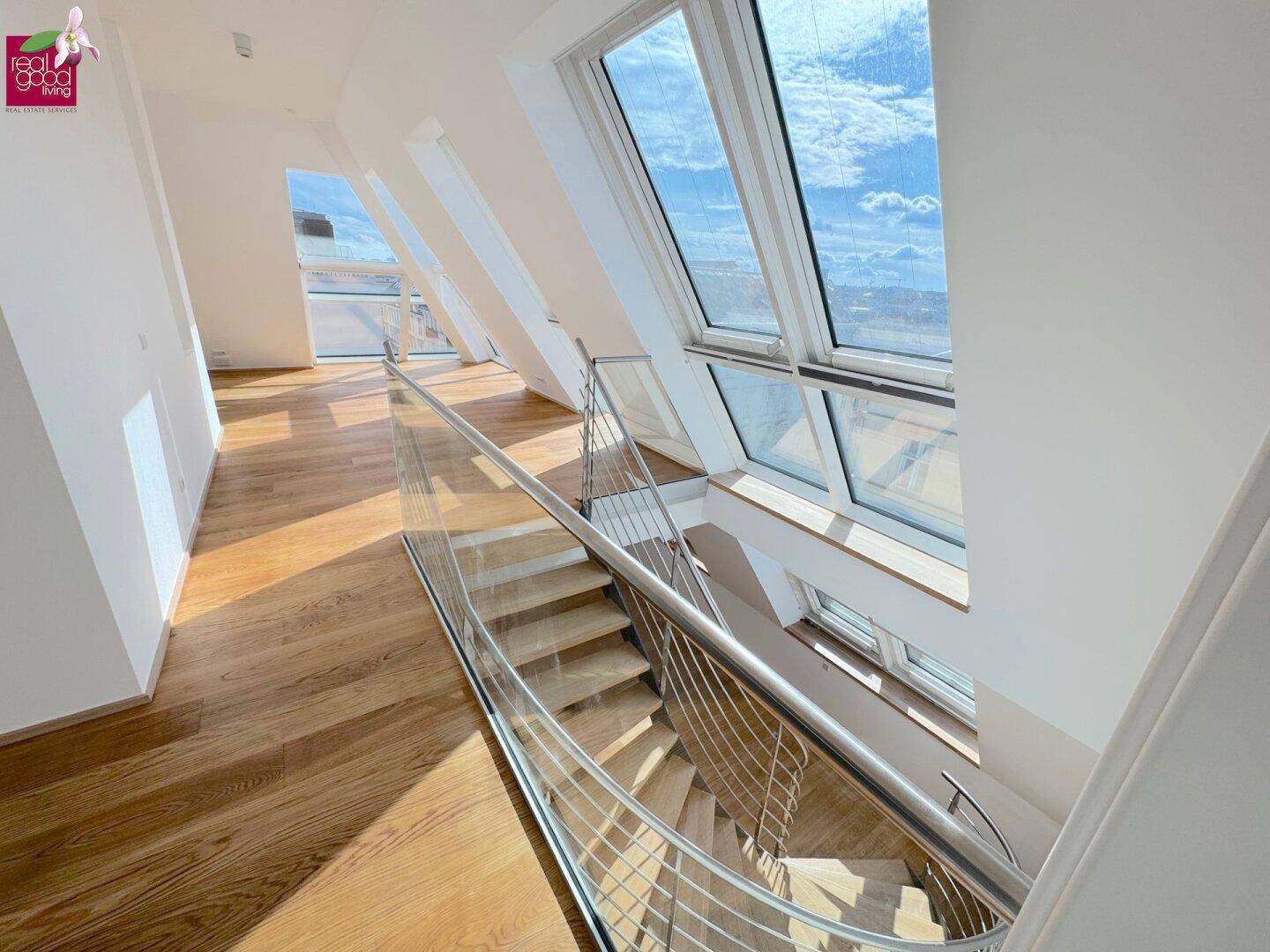 Moderne Treppe mit Glasgeländer und Holzstufen, umgeben von großen Dachfenstern mit Himmelsblick.