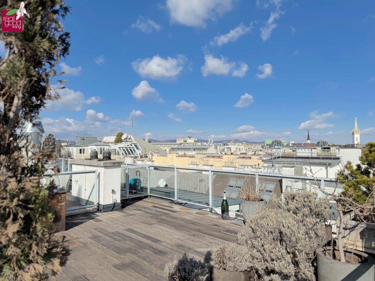 Geräumige Dachterrasse mit Holzbelag, Glasgeländer und Stadtblick unter blauem Himmel.