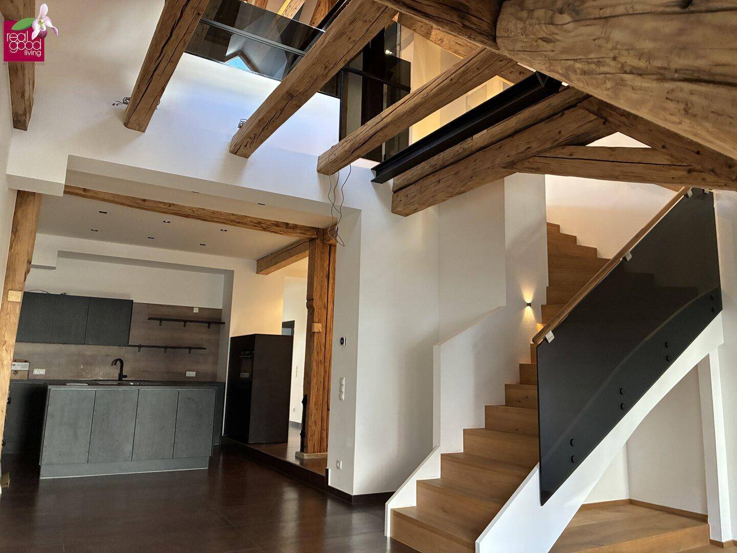 Offener Wohnbereich mit moderner Küche, Holztreppe und sichtbaren Holzbalken.