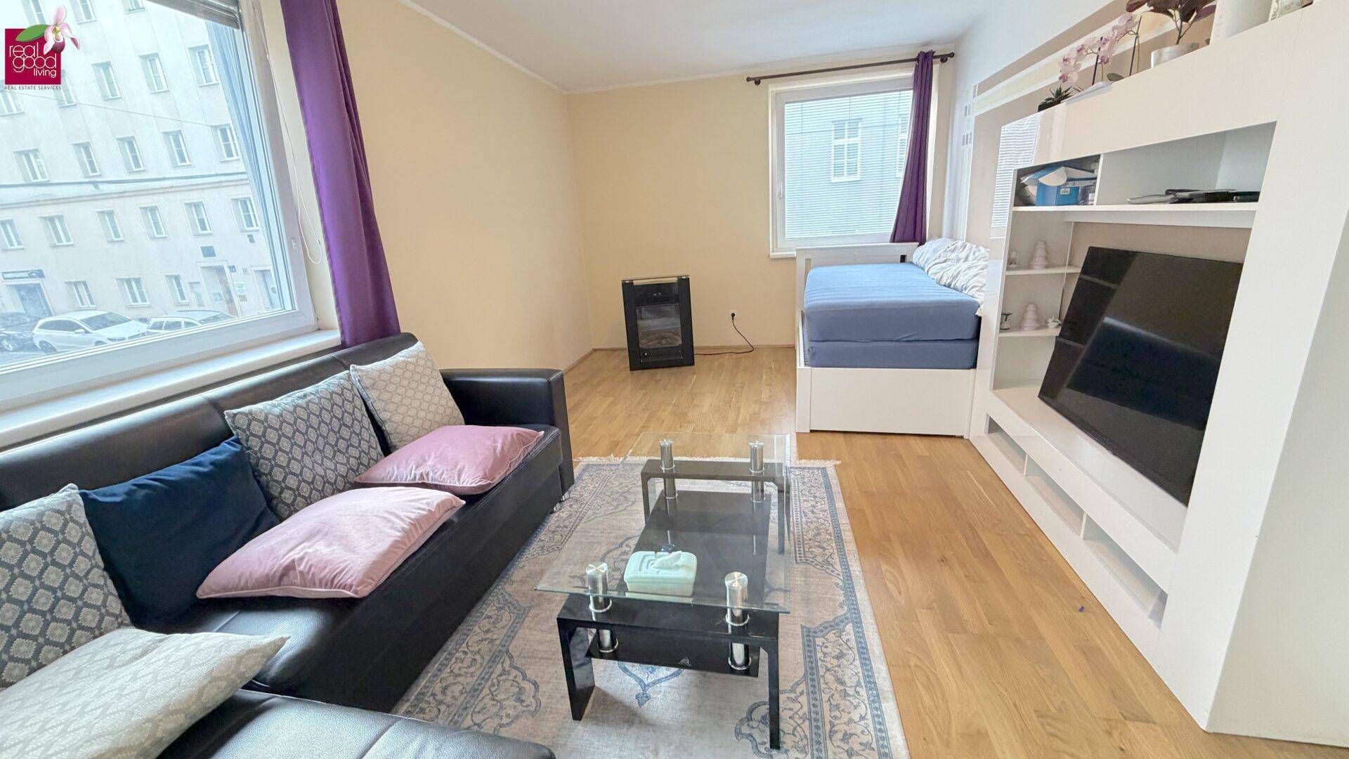 Wohnzimmer mit Sofa, Couchtisch, TV-Möbel und einem integrierten Bettbereich, hell durch große Fenster.