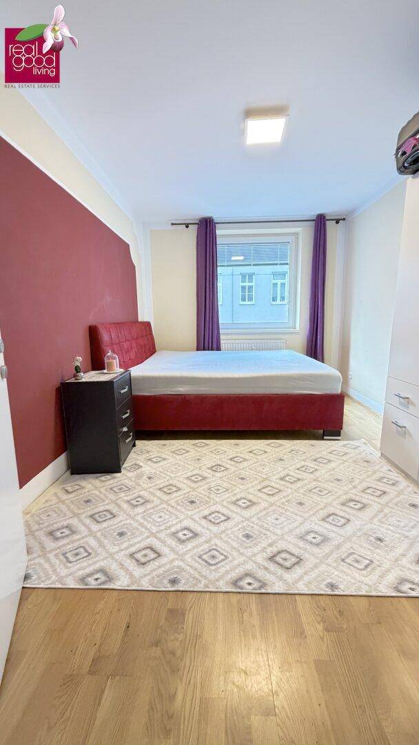 Schlafzimmer mit rotem Bett, Holzfußboden und einem Fenster, das für natürliches Licht sorgt.