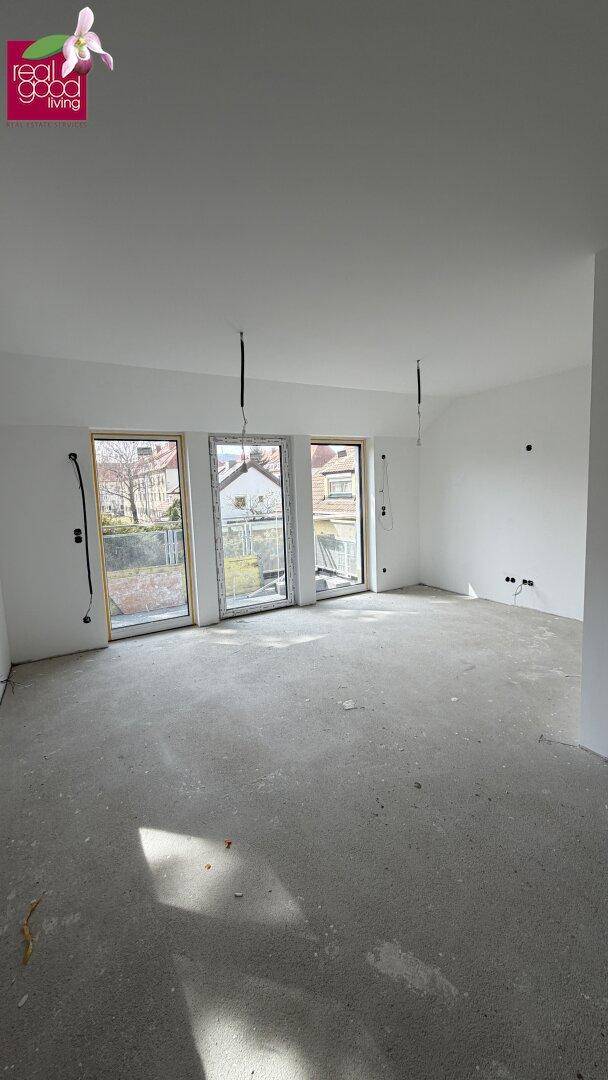 Heller Innenraum im Rohbau mit großen Fenstern und Betonboden, bereit für den Ausbau.
