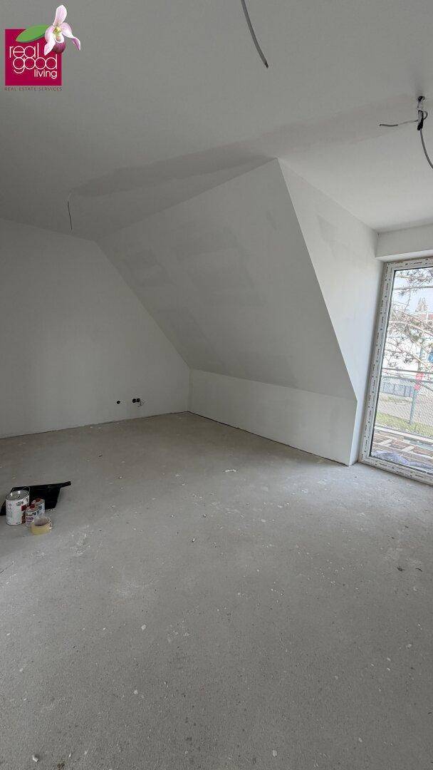 Unmöblierter Raum im Dachgeschoss mit schräger Decke, großem Fenster und Betonboden.