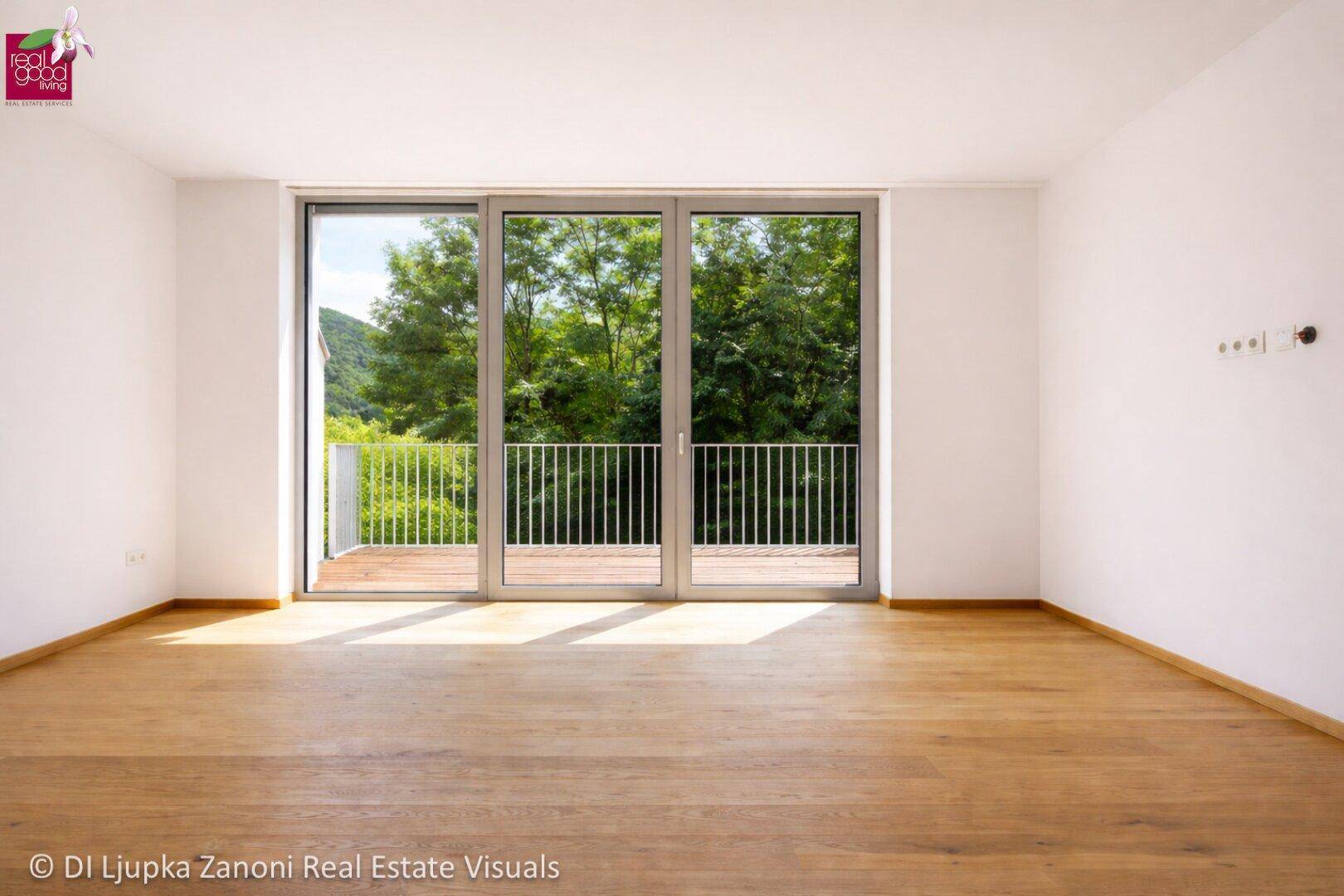Heller Raum mit Holzboden und Zugang zu einem Balkon mit Blick ins Grüne.