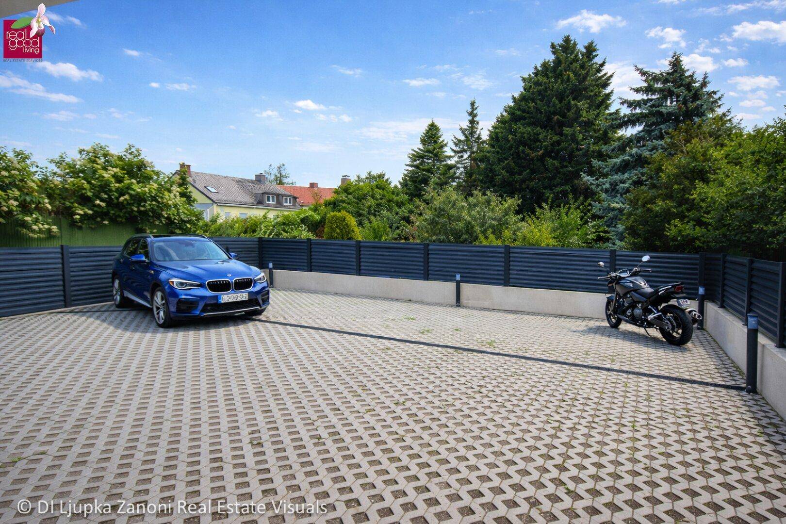 Geräumiger Außenparkplatz mit Pflastersteinen, umgeben von einem Zaun und Bäumen, mit einem Auto und Motorrad.