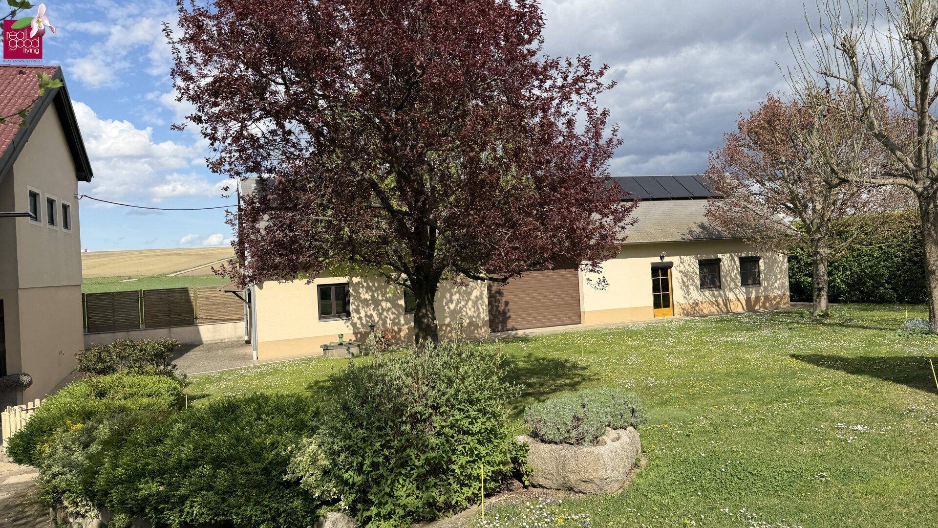 Weitläufiger Gartenbereich mit Bäumen und Sträuchern, angrenzend an ein Gebäude mit Garagentor.