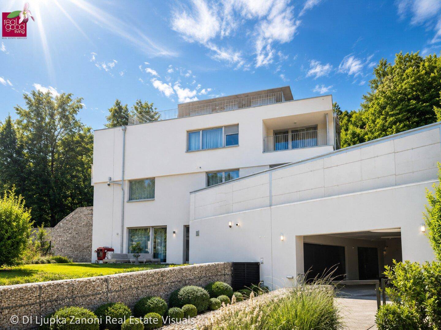 Moderne weiße Villa mit großen Fenstern, Balkonen und integrierter Garage in grüner Umgebung.