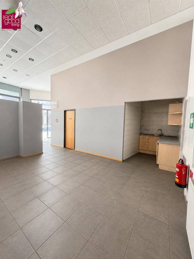 Geräumiger Gewerberaum mit Fliesenboden, hohen Decken und einer kleinen Küchenzeile im Hintergrund.