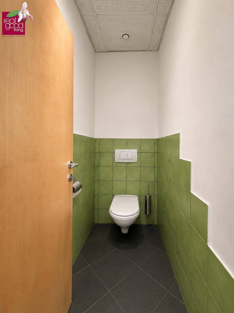 Separate Toilette mit grün gefliesten Wänden, weißer Keramik und dunklem Fliesenboden.