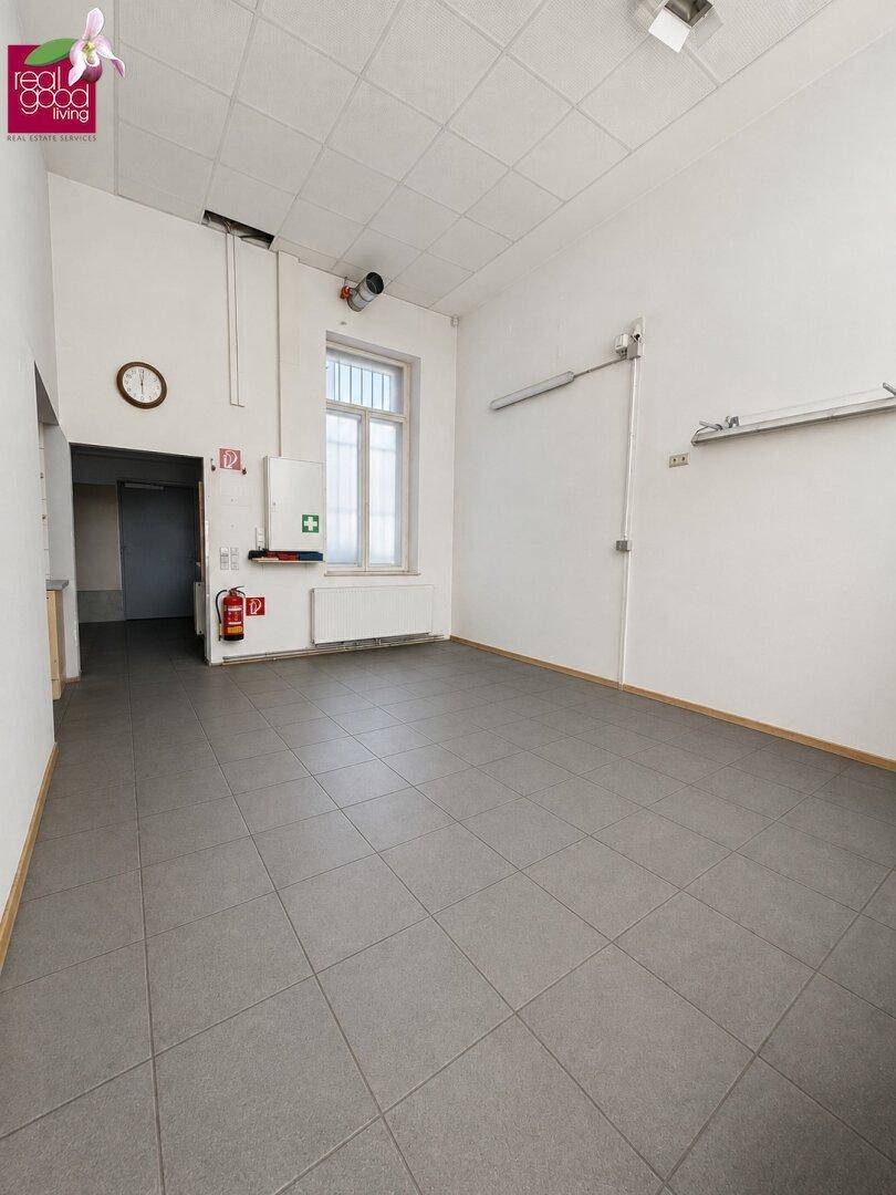 Großer, leerer Gewerberaum mit Fliesenboden, hohen Decken und einem Fenster zur Straße.