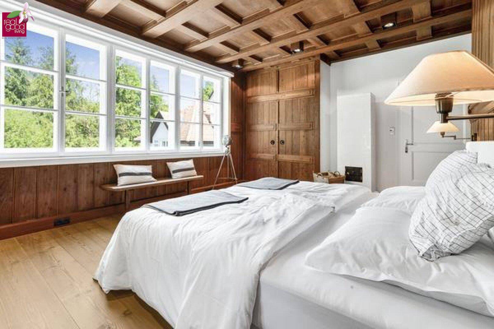 Gemütliches Schlafzimmer mit Holzbalkendecke, großen Fenstern, Holzwänden und Blick ins Grüne.
