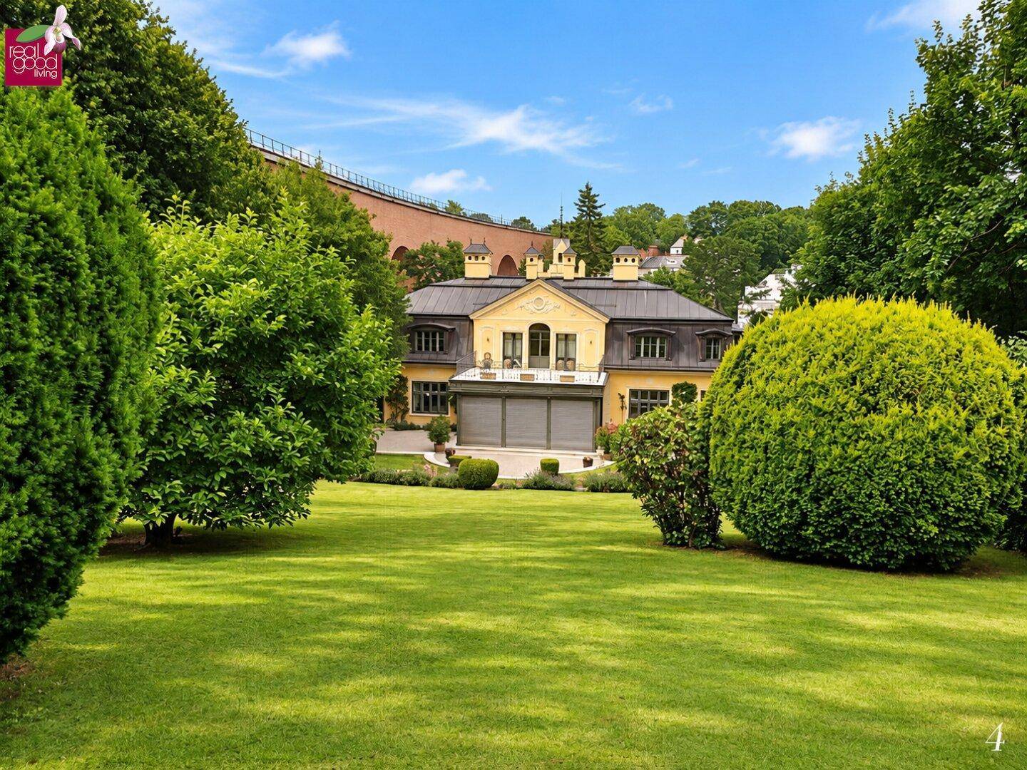 Weitläufiger Blick auf die gelbe Villa von einem großen, gepflegten Rasenbereich mit üppiger Bepflanzung.