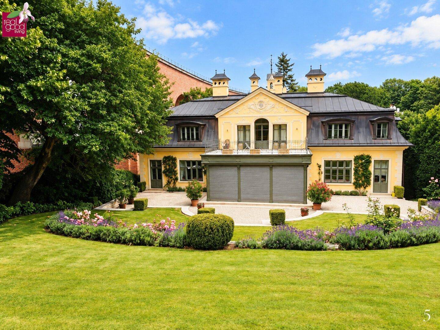 Rückansicht der gelben Villa mit kleinem Balkon, umgeben von blühenden Blumenbeeten und einem weitläufigen Rasen.