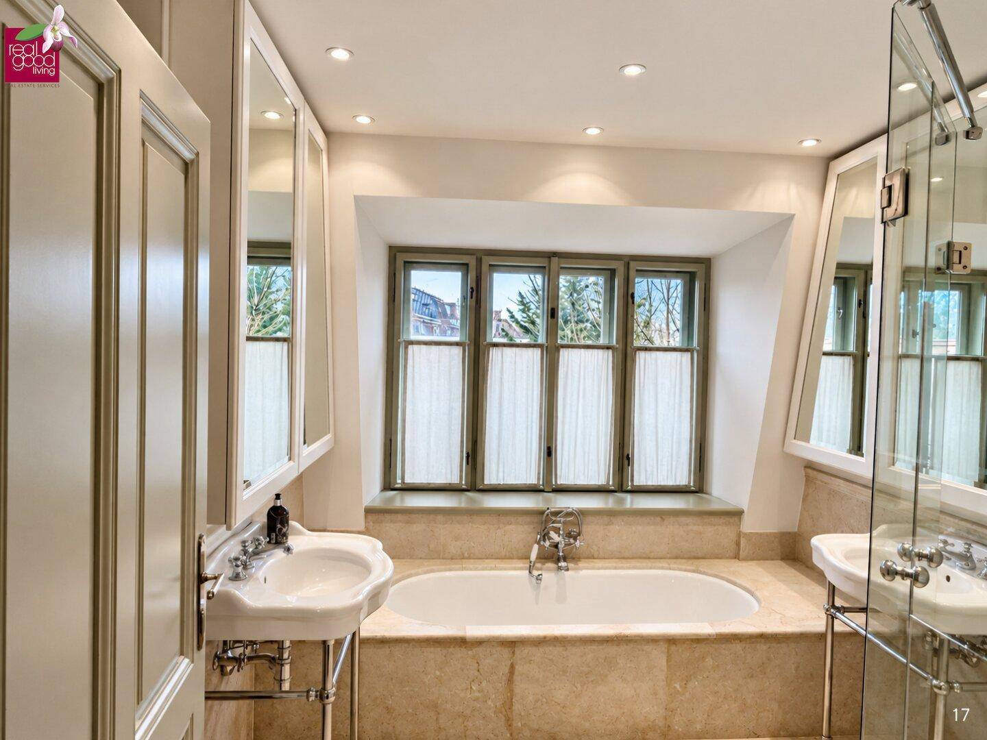 Helles Badezimmer mit eingebauter Badewanne, zwei Waschbecken und einem großen Fenster, in hellem Marmor gehalten.
