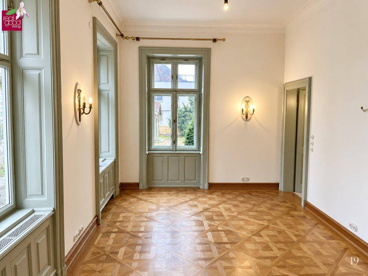 Heller Raum mit gemustertem Parkettboden, weißen Wänden und hohen Fenstern mit grünen Rahmen.