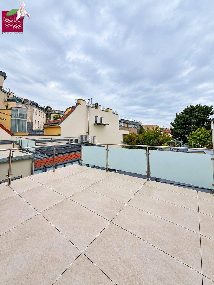 Große Dachterrasse mit Fliesenboden und Glasgeländer, umgeben von städtischer Bebauung.